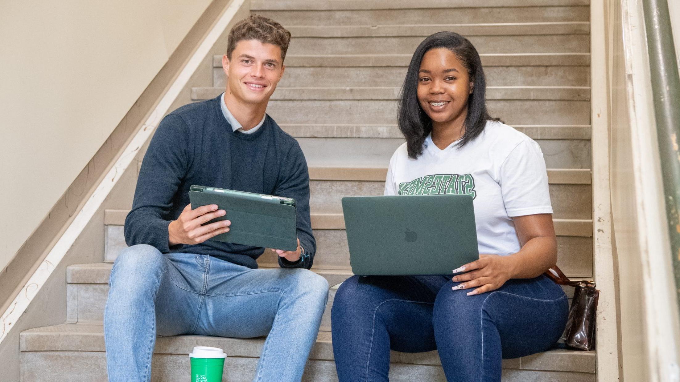 两个学生，一个带着笔记本电脑，一个带着ipad，坐在楼梯上微笑.
