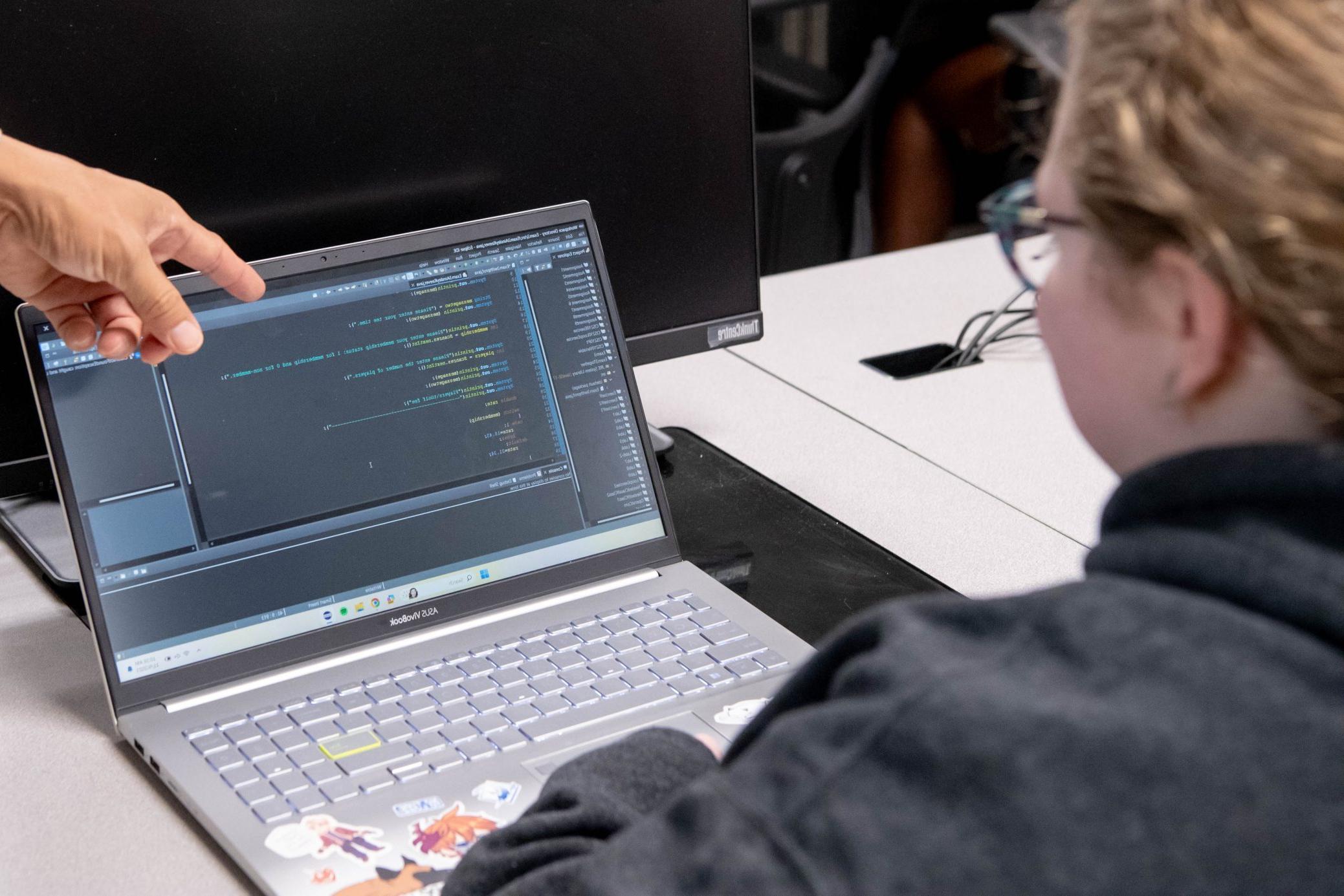学生坐在笔记本电脑前，教授用手指着电脑屏幕上的代码.